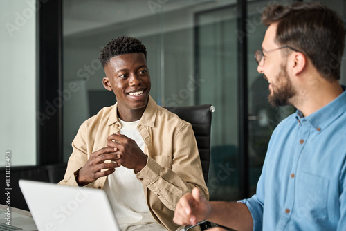 Two busy diverse male employees using laptop computer talking in office. Professional young business men workers team having conversation working on project sharing ideas sitting at work desk.