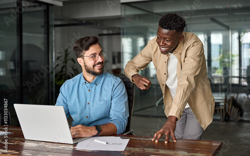 Happy Latin team leader teaching young male African coworker explaining online project strategy. Two busy diverse professional software engineers working in teamwork talking at work desk with laptop.