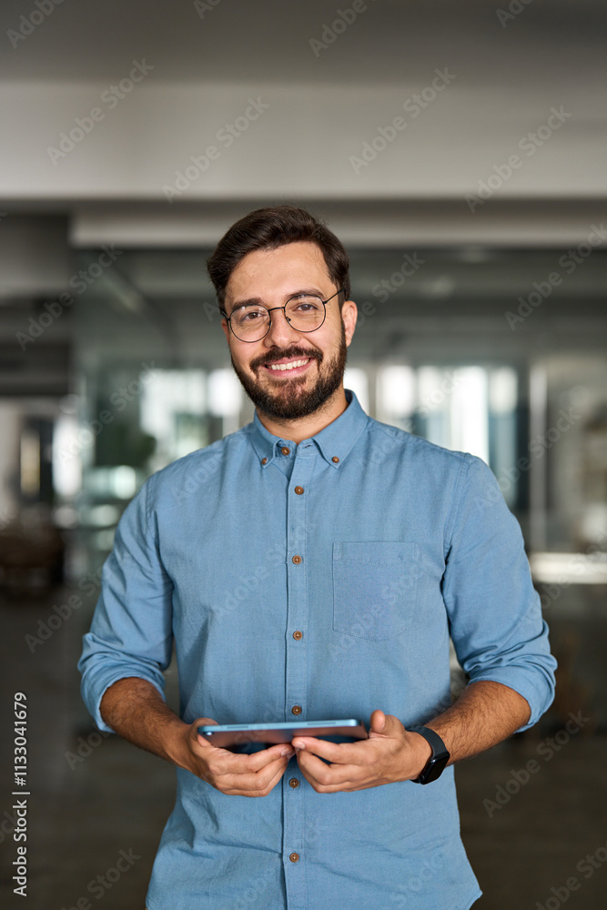 Obraz premium Happy young professional Latin business man looking at camera standing at work using tablet. Portrait of smiling male company worker employee, project manager or entrepreneur in office.