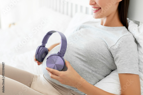 European pregnant woman holding wireless headphones near belly, lady using earphones for playing music to baby in womb, sitting on bed, cropped photo