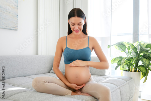 Happy young pregnant woman sitting on couch in living room, tenderly embracing belly, looking at her baby bump with love Free Download Happy young pregnant woman sitting on couch in living room, tenderly embracing belly, looking at her baby bump with love - GFXLoad