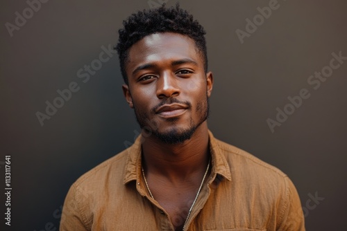 A young Black man with a short, textured hairstyle, wearing a brown shirt, gazes directly at the camera with a calm expression.