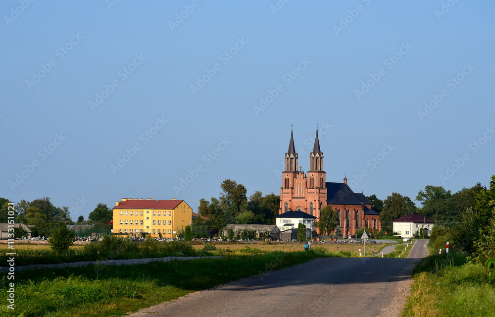 Obraz premium A close up on an asphalt road leading to a small town or village with a towering, big church or chapel made out of red brick located next to vast fields used for agriculture and cattle grazing