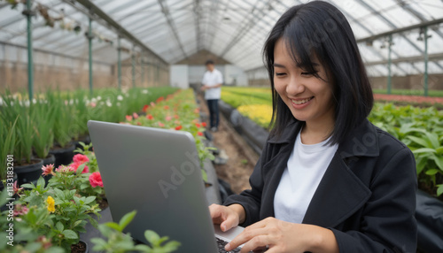 Asian woman with shoulder length black hair, in a black coat, smiling while working on a laptop in a greenhouse photo