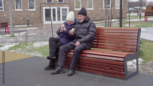 Elderly couple in warm winter clothing sitting on a bench, enjoying ice cream cones outdoors on a cold day. Retirement life concept