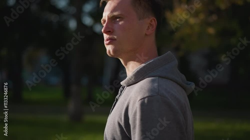 Young man with short dark brown hair wearing light gray hoodie, serious and contemplative expression while looking into distance, with blurred green and yellow-brown foliage in background