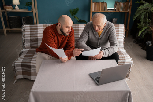 Middle aged son helping his father with paperwork and paying bills online while sitting in the living room at home photo