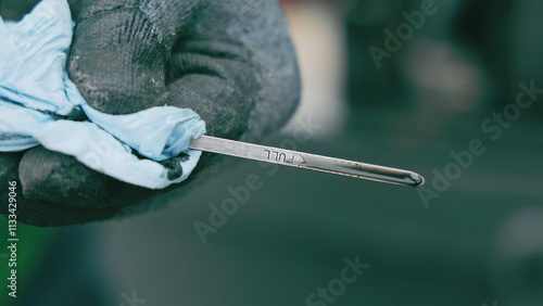 Checking Oil Level with Dipstick. A close-up view of a gloved hand holding an oil dipstick marked with the word FULL, while wiping it clean with a blue cloth, against a blurred background.