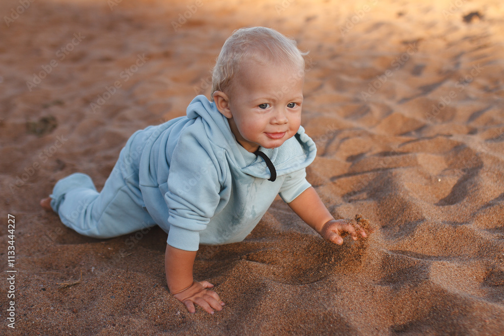 Fototapeta premium Happy toddler in blue hoodie crawls along beach in sand on sunny day