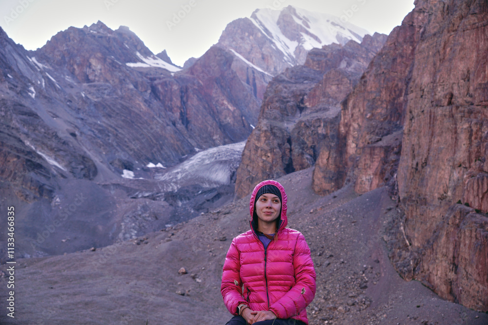 Fototapeta premium A young girl in bright clothes is sitting on a rock and smiling in the mountains against the background of rocks and snowy peaks. Trekking in the mountains in summer