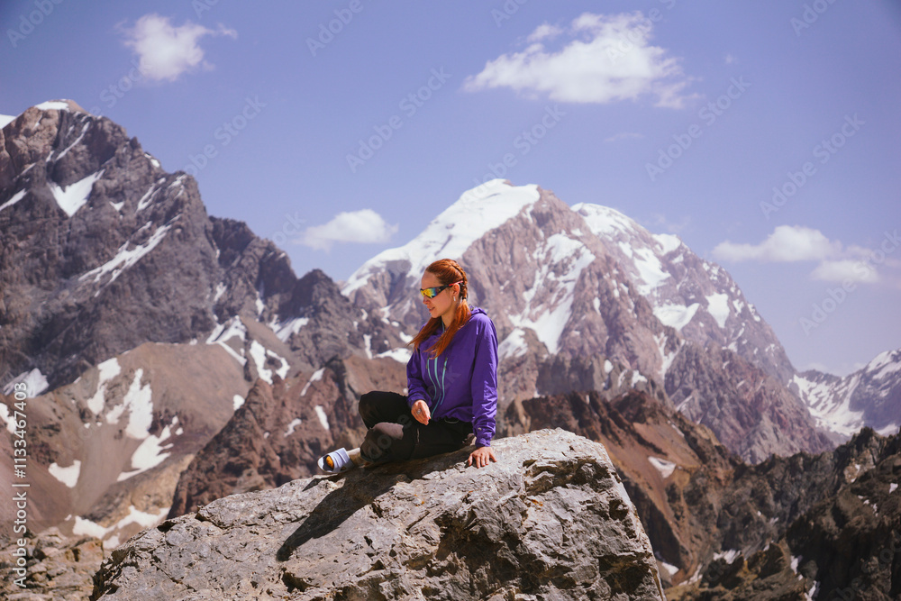 Fototapeta premium A young girl in bright clothes is sitting on a rock and smiling in the mountains against the background of rocks and snowy peaks. Trekking in the mountains in summer