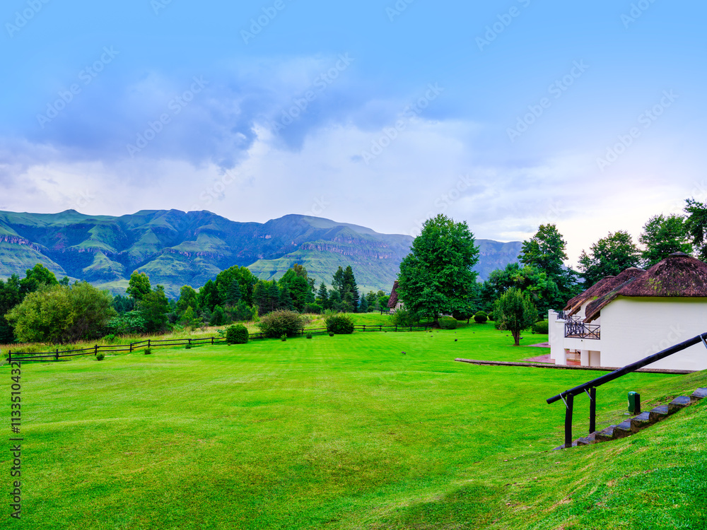 Lush grass field with a thatch-roof house and Drakensberg mountain in the background, KwaZulu-Natal, South Africa