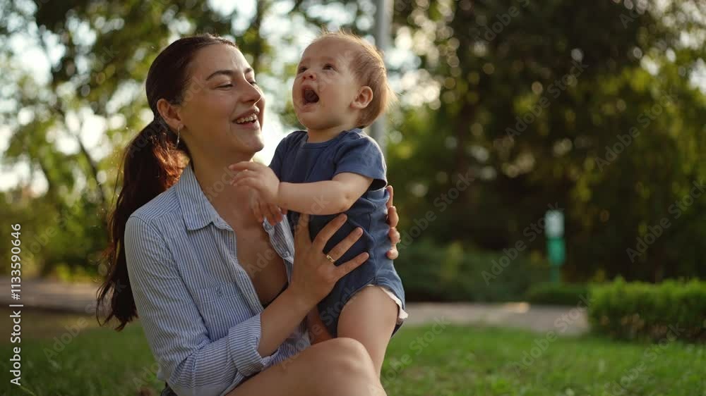 Happy brunette woman in a jacket holds her active little baby daughter in her arms and rejoices at her during her vacation in the park in summer Happy brunette woman in a jacket holds her active little baby daughter in her arms and rejoices at her during her vacation in the park in summer