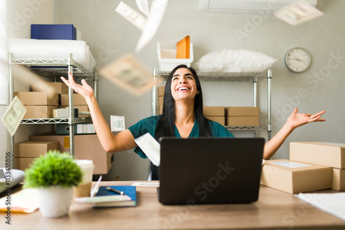 Excited businesswoman throwing money in the air while working on laptop in her warehouse, celebrating success of her small business photo