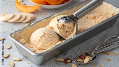 A sleek silver s emerges from a lidless container of soft peach ice cream mingling with a few lightly toasted almond slices that rest on the surface. The scoop glistens with photo