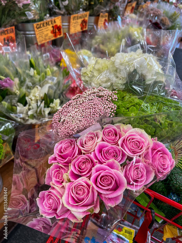 A vibrant selection of pink roses, white hydrangeas, green foliage, and pink filler flowers wrapped in plastic, displayed in a shopping cart at a colorful flower market.