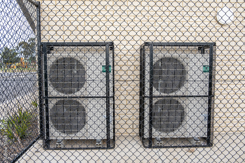Schilderij op canvas Large commercial air conditioning units, enclosed in protective metal cages, are installed against a wall as an anti-theft measure