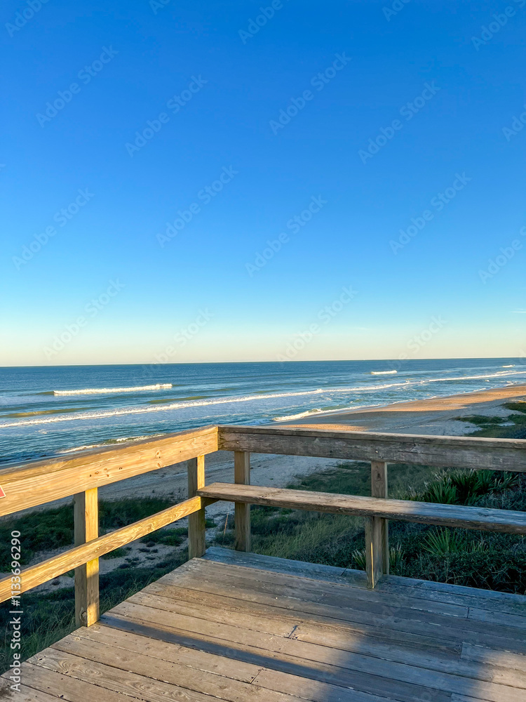 Obraz premium A wooden beach lookout overlooks serene ocean waves, a golden sandy shore, and lush dunes under a clear blue sky, capturing a peaceful coastal escape.