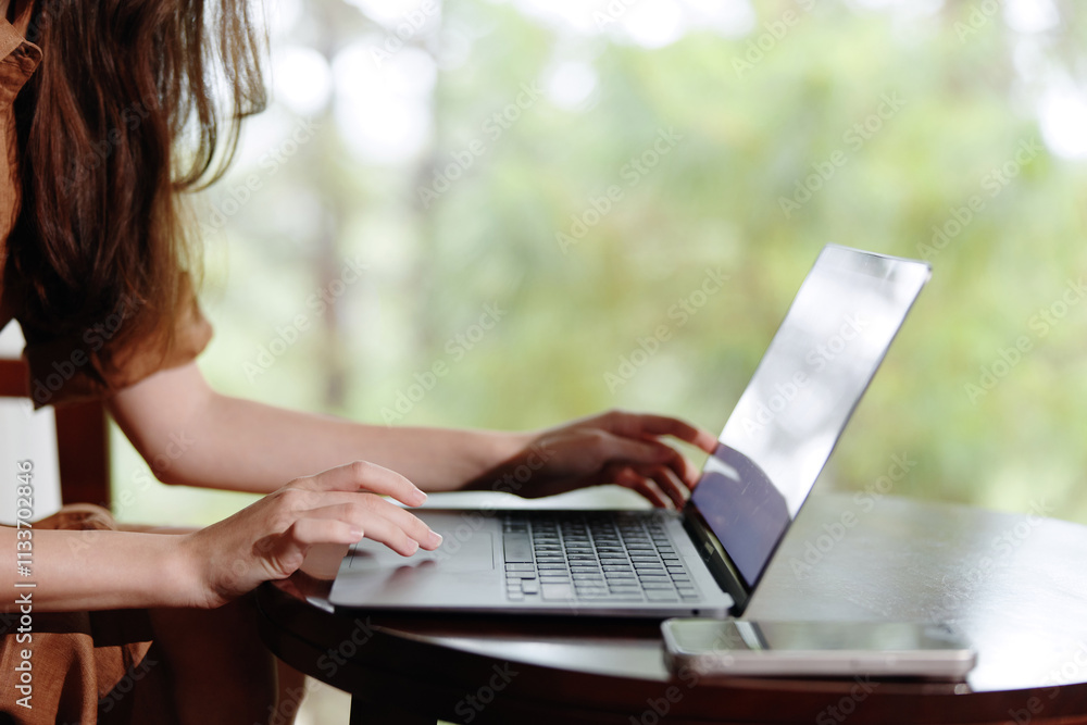 A woman working on a laptop at a wooden table, showcasing focus and productivity in a tranquil outdoor setting with blurred greenery in the background A woman working on a laptop at a wooden table, showcasing focus and productivity in a tranquil outdoor setting with blurred greenery in the background