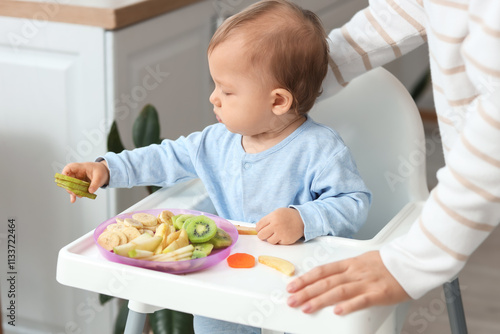 Cute little baby with fruits in high chair and mother at home