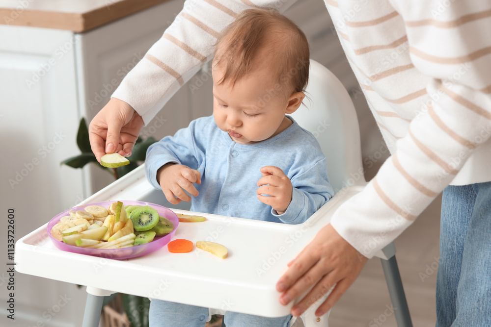 Mother feeding her little baby with fruits in high chair at home Mother feeding her little baby with fruits in high chair at home
