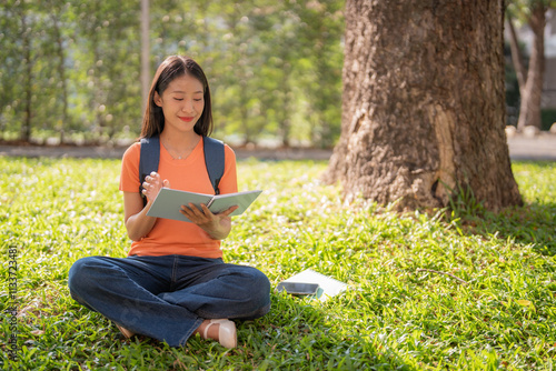 Female college student sitting under a tree on the grass in a university campus park, studying and taking notes while enjoying the pleasant weather and sunshine