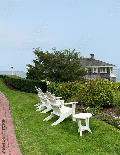 White Adirondack chairs and a side table sit along a manicured lawn with a view of a coastal house, hedges, and blooming flowers on a clear day by the sea.