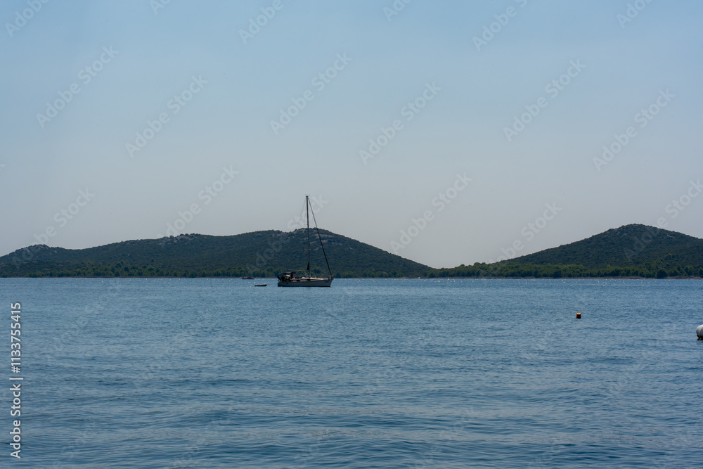 Serene sailboat floating on calm blue sea with lush green hills in the background under clear sky on a sunny day. Concept of tranquility, nature, and nautical adventure. Serene sailboat floating on calm blue sea with lush green hills in the background under clear sky on a sunny day. Concept of tranquility, nature, and nautical adventure.