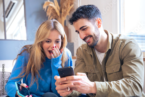 Bilde på lerret A young couple seated indoors sharing a surprising and joyful moment while looking at a smartphone