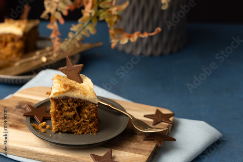 Iced carrot cake sllice served on grey pewter plate with chocolate star and mid blue table cloth, lplant leaves in background in soft focus uxury earthy feel photo