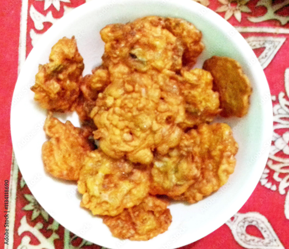 A bowl filled with golden, crispy fritters is placed on a red patterned tablecloth. The fritters appear to be deep-fried, offering a delicious and savory snack option. A bowl filled with golden, crispy fritters is placed on a red patterned tablecloth. The fritters appear to be deep-fried, offering a delicious and savory snack option.