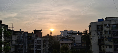 The sun sets over a cityscape marked by clustered apartment buildings, casting a warm glow across the sky. Clouds add texture to the serene scene while greenery peeks out between structures. photo