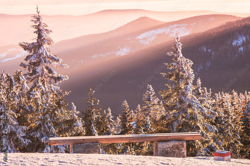 Eastern Sudetes winter landscape, wooden bench on mountain hiking trail with view of mountain range, sun rays illuminate mountain slopes in evening.