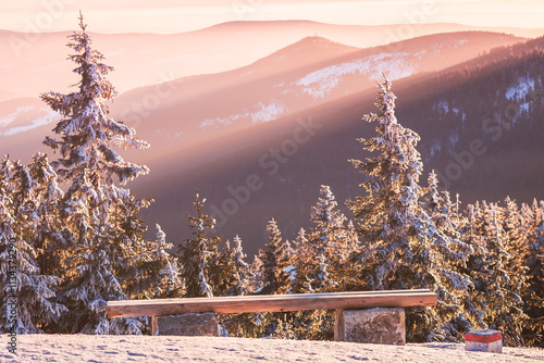 Obraz na płótnie Eastern Sudetes winter landscape, wooden bench on mountain hiking trail with view of mountain range, sun rays illuminate mountain slopes in evening. w salonie
