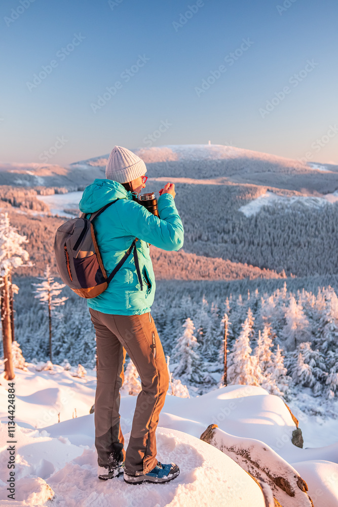 Eastern Sudetes, Young woman hikes on a snow-covered mountain trail, stands on a viewpoint and eats a warm meal, winter landscape in the mountains at sunset.