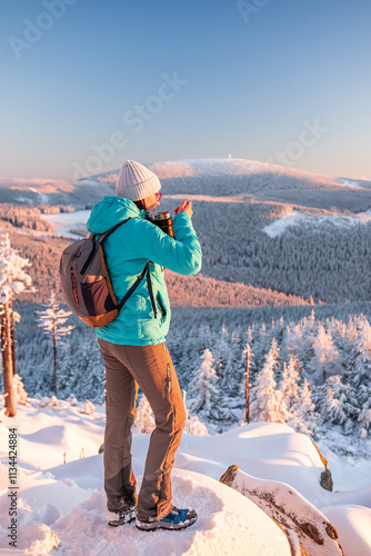 Eastern Sudetes, Young woman hikes on a snow-covered mountain trail, stands on a viewpoint and eats a warm meal, winter landscape in the mountains at sunset.