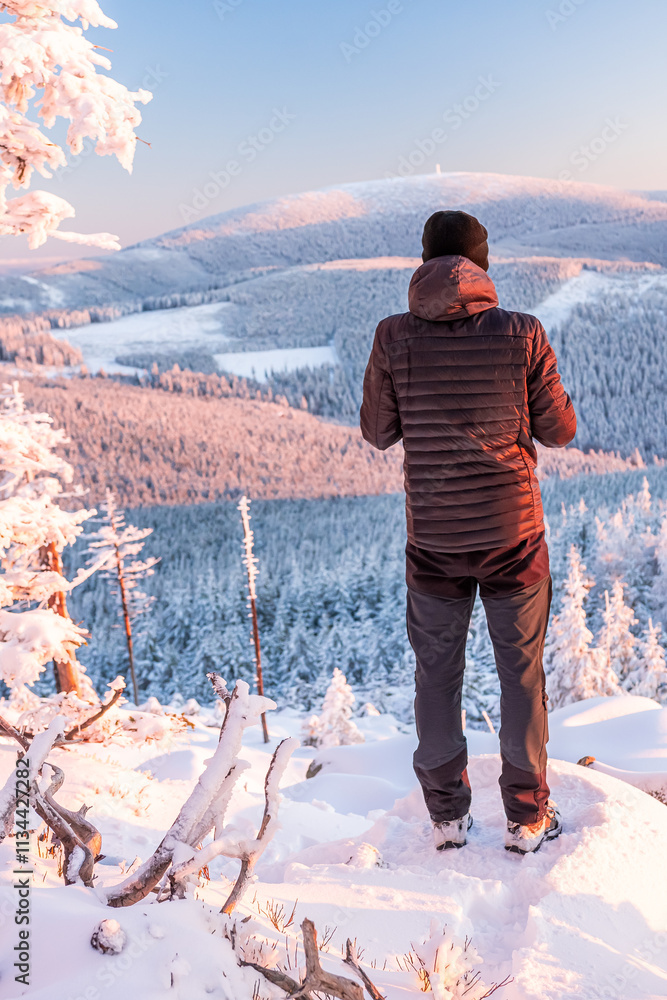 Eastern Sudetes, A young man hikes along a snow-covered mountain trail, stands on a viewpoint and looks at the snow-capped peaks and forest, winter landscape in the mountains.