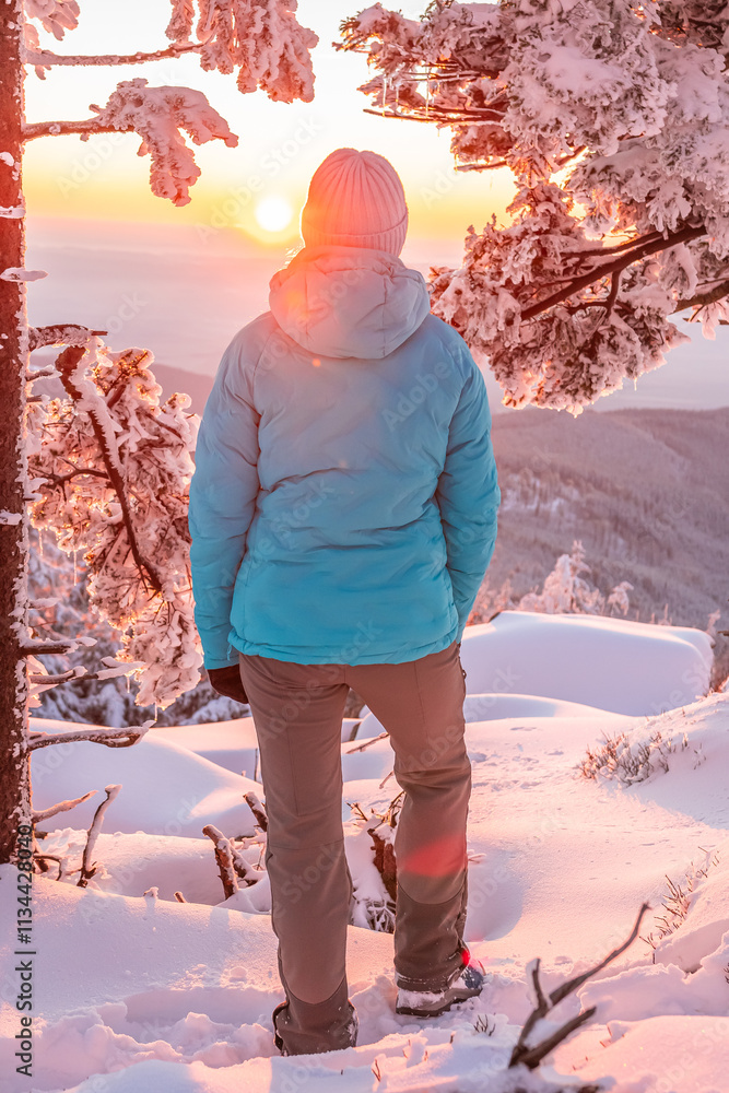 Eastern Sudetes, Young woman hikes along a snow-covered mountain trail, stands on a viewpoint and looks at the sunset, winter landscape in the mountains.