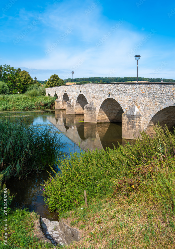 Fototapeta premium Stone bridge with arches over Wornitz river in Germany, surrounded by lush greenery.