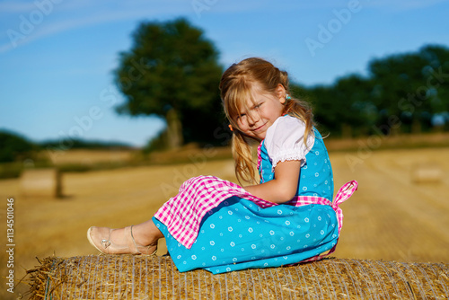 Cute little kid girl in traditional Bavarian costume in wheat field. Happy child with hay bale during Oktoberfest in Munich. Preschool girl play at hay bales during summer harvest time in Germany. photo