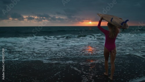 Woman surfer carries a surfboard, waves on the shore or beach at dawn. A beautiful girl goes surfing on the island of Bali. A female surfer in a red swimsuit holds a board for the sea. morning sport