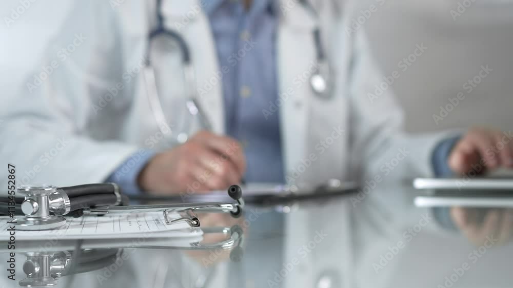 Stethoscope and medication history records form are on a glass table before a male doctor working with laptop in clinical office background. Medicine concept Stethoscope and medication history records form are on a glass table before a male doctor working with laptop in clinical office background. Medicine concept