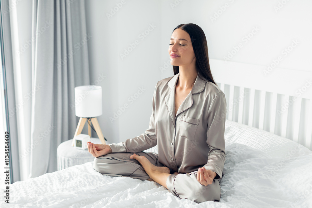 Serene young woman sitting cross-legged on white bed, with her eyes closed and hands in meditative pose Serene young woman sitting cross-legged on white bed, with her eyes closed and hands in meditative pose