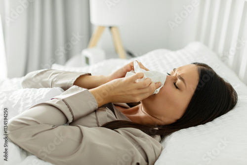 Sick lady suffering from running stuffy nose, lying in bed and blowing her nose using paper napkin tissue. Cold and flu concept