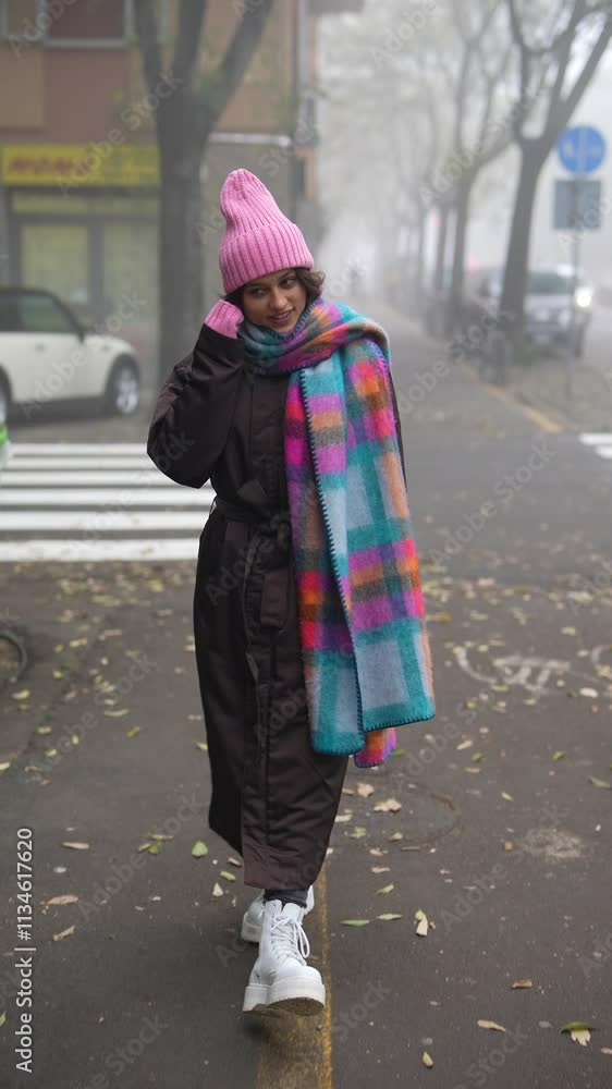 A woman walks gracefully in a foggy street while wearing a colorful scarf and a stylish hat A woman walks gracefully in a foggy street while wearing a colorful scarf and a stylish hat