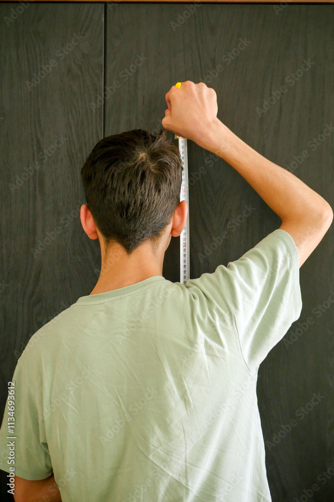 A young man trying to measure his height with a tape measure