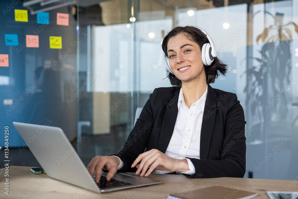 Businesswoman in office wearing headphones working on laptop, smiling at desk. Engaged in virtual meeting, focused on work, technology, communication, productivity. Modern and confident atmosphere. Businesswoman in office wearing headphones working on laptop, smiling at desk. Engaged in virtual meeting, focused on work, technology, communication, productivity. Modern and confident atmosphere.