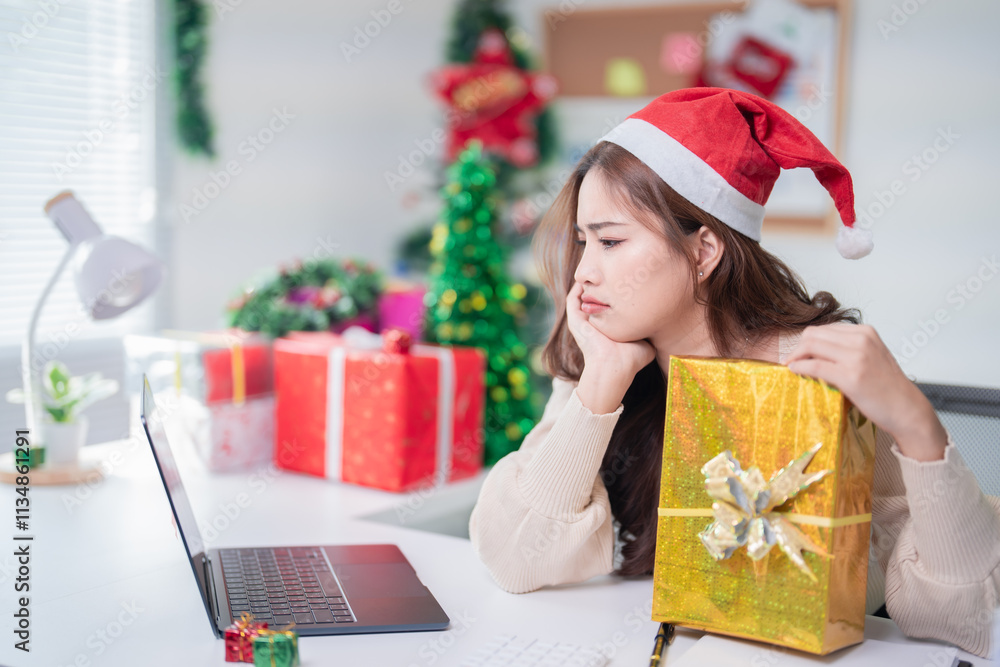 Bored young asian office worker wearing santa hat is holding a christmas present and looking sadly at her laptop during online christmas party Bored young asian office worker wearing santa hat is holding a christmas present and looking sadly at her laptop during online christmas party