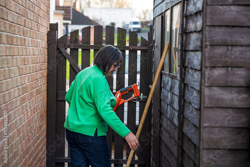 Woman wearing Green Top Storing Weed Whacker Strimmer and Garden Broom in Shed photo