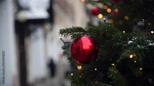 Close-up view of red Christmas bauble hanging on green spruce tree on winter city street. Blurred people walking on sidewalk in the background. Handheld video. Soft focus. Holidays theme.